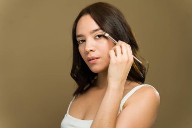 Genç bir kadın bir fırçayla göz farı sürüyor, stüdyo ortamında doğal güzelliğini geliştiriyor.
