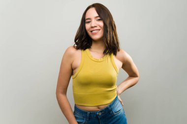 Elini kalçasında gezdiren, kendine güveni ve günlük tarzıyla neşeli genç bir kadının stüdyo portresi.