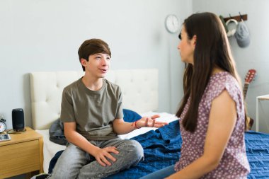 Latin kökenli genç bir çocuk, odasında oturmuş elleriyle el kol hareketi yaparken annesiyle sohbet ediyor.