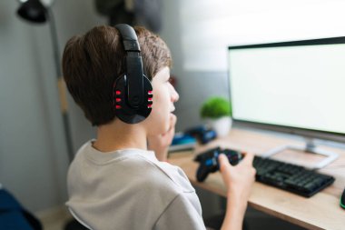 Kulaklık takan ve bilgisayarında video oyunu oynayan bir çocuk yatak odasında bir kumanda kullanıyor.