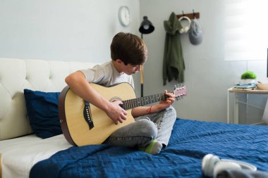 Genç müzisyen yatak odasında akustik gitar çalışıyor, evde hobisinin tadını çıkarıyor.