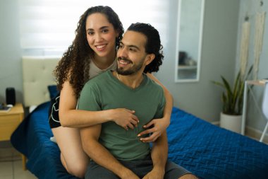 Latin çift gülümsüyor ve modern yatak odasında yatakta kucaklaşıyorlar, samimiyet ve birliktelikten zevk alıyorlar.