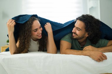 Sıcak battaniyelerinin altında rahatlayan, gülümseyen ve sıcak yatak odalarında neşeli yakınlık anlarını paylaşan Latin çifti.