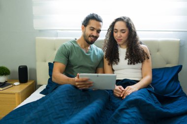 Genç İspanyol çift yatakta dinleniyor, rahat yatak odalarında birlikte bir tabletle film izliyorlar, rahatlık ve bağ kuruyorlar.
