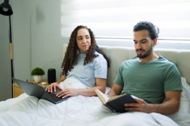 Rahat İspanyol çift, erkek arkadaşı kitap okurken dizüstü bilgisayarıyla uzaktan çalışan kadınla yatakta vakit geçiriyor.
