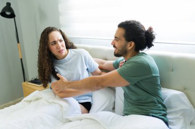 Genç çift yatakta, erkek arkadaşı partnerine sarılmaya çalışırken kadın şüpheli bir ifadeyle uzaklaşıyor.