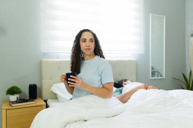 Genç bir kadın yatakta sıcak bir fincan kahvenin tadını çıkarıyor. Ortağı yanında mışıl mışıl uyurken huzurlu bir sabahın tadını çıkarıyor.