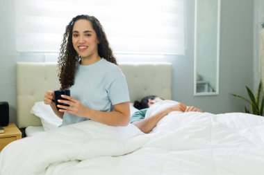 Bir kadın, rahat bir yatak odasında, arka planda yatağında yatarken elinde kupa ve gülümsüyor.