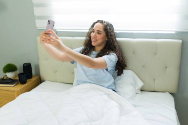 Neşeli İspanyol kadın sabah yatakta rahat bir şekilde otururken akıllı telefonuyla selfie çekiyor.