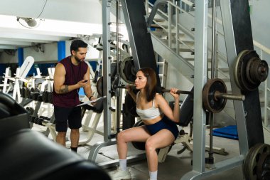 Güzel bir kadın spor salonunda halterle çömeliyor, kişisel antrenörüyle gelişimi izliyor ve takip ediyor.