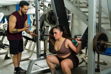 Spor salonunda bir halterle çömelme egzersizi yapan bir kadın, erkek özel antrenörden rehberlik alıyor.