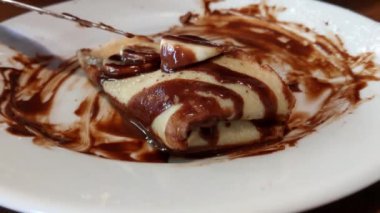 The girl cuts a sweet pancake with chocolate and banana with a knife