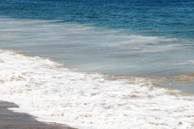 Günlük kıyafetler giyen bir adam okyanusun yanındaki kumlu sahil boyunca yürüyor. Dalgalar yavaşça sahile vuruyor ellerini cebinde gezdirirken..