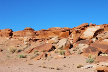 Büyük bir kaya oluşumu açık mavi bir gökyüzünün altında çorak bir çöl arazisinin ortasında belirgin bir şekilde duruyor. Kaya oluşumu dayanıklı ve yıpranmış. Jeolojik süreçlerin ham gücünü gösteriyor..
