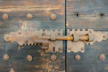 Yıpranmış ahşap bir kapının üzerinde antika bir cıvata kilidi, kırsal cazibe ve klasik tasarım sergiliyor. Metal kilit, karmaşık detayları ve eskimiş dokusuyla, tarihsel hissi artırır, onu güvenlik, mimari, bir