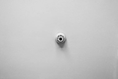 Minimalist black and white image featuring a smoke detector mounted on a white wall. represents themes of safety, security, and modern home design elements. ideal for use in articles, brochures, or websites focused on home safety and security systems