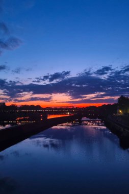 Alacakaranlıkta sakin nehir manzarası, dramatik bulut oluşumlarıyla canlı bir günbatımı. Sakin sulardaki yansımalar, zıt sıcak ve serin tonların altını çiziyor. Duvar sanatı ve seyahat posterleri için huzurlu ama çarpıcı bir görsel yaratıyor..