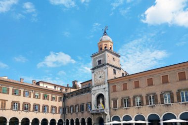 Modena, İtalya, 12 Mayıs 2024: Parlak mavi gökyüzünün altında önde gelen bir saat kulesinin yer aldığı tarihi bir kasaba meydanı. Meydan, birden fazla penceresi olan klasik, iyi korunmuş binalarla çevrili ve mimari güzellik ve tarih sahneleri yaratıyor.