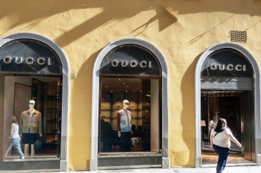 Florence, Italia, 10 Mayıs 2024: Gucci mağaza vitrininde, büyük cam pencerelerin arkasında şık kıyafetler giymiş iki manken. Parlak, güneşli kentsel ortam, yoldan geçenlerin canlılık katmasıyla lüks ortamı geliştirir.