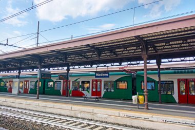 Modena, İtalya, 12 Mayıs 2024: Modena, İtalya 'daki platformda kırmızı ve yeşil üniformalı bir trenin bulunduğu modern tren istasyonu. Kentsel mimari tasarımı vurgulayarak, kablolar ve tabelalar görülebilir durumda. Avrupa taşımacılığının sahne özü
