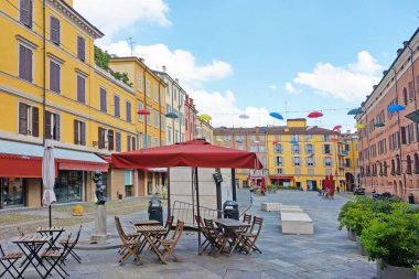 Modena, İtalya, 12 Mayıs 2024: Canlı binaları ve asılı şemsiyelerle süslenmiş bir İtalyan kasabasında parlak ve neşeli bir meydan. Açık hava kafe masaları ve sandalyeler misafirperver bir atmosfer sağlar ve canlı renkler canlı ambiyansı, kimliği vurgular.