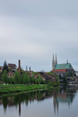 Gorlitz, Almanya, 13 Nisan 2024: Riverside kasabası sakin sulara yansıyan uzun ikiz kuleleri olan tarihi bir kilise. Kanolar nehri dolduruyor, yemyeşil ağaçlar ve arka planda büyüleyici binalar bulutlu bir gökyüzünün altında..