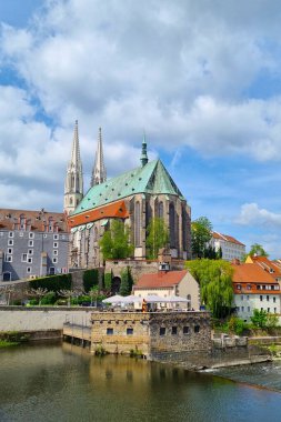 Gorlitz, Almanya, 13 Nisan 2024: Büyüleyici bir köyde sakin bir nehir kıyısında ikiz kuleleri olan güzel bir katedral. Kırmızı çatılı binalar ve yemyeşil yeşillikler mimari harikaların etrafını sarar. Parçalı bulutlu bir gökyüzünün arka planına kurulur.