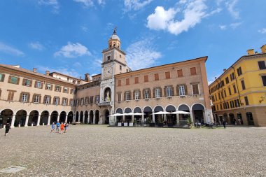 Modena, İtalya, 12 Mayıs 2024: Klasik Avrupa mimarisi ve canlı binalarla çevrili tarihi bir saat kulesinin yer aldığı Picturesque şehir meydanı. Turistler açık bir yaz gökyüzü altında büyüleyici atmosferin tadını çıkararak bölgeyi keşfediyorlar.