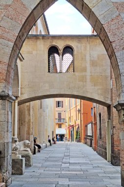 Modena, İtalya, 12 Mayıs 2024: Tarihi kemerli mimarinin yer aldığı ortaçağ İtalyan geçidi. Dar cadde antik taş binalarla dolu ve birkaç kişi rahatça banklarda oturuyor. Pastel renkli binalar ve bir temizlik.