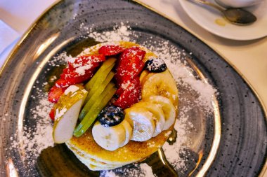 Üzerine taze çilek, kivi dilimleri, muz parçaları ve yaban mersini serpiştirilmiş sanatsal olarak pudra şekeri serpiştirilmiş altın krep yığını. Kahvaltı temalı menüler, mutfak blogları ve yemek fotoğrafçılığı projeleri için mükemmel..