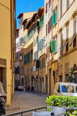 Bolonya, İtalya, 19 Mayıs 2024: Canlı yeşil kepenkler ve geleneksel mimarinin yer aldığı tarihi binalarla dolu dar bir Avrupa caddesi. Yayalar kaldırım taşı yolu boyunca yürür ve güneşli, bulutsuz bir gökyüzü sıcak atmosferi güçlendirir, anne.