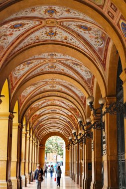 Bolonya, İtalya, 18 Mayıs 2024: Güzel dekore edilmiş tavanlar ve sütunlar içeren zarif kemer yolu. Dört kişi yürüyor, ölçek ve şehir hayatı hissi ekliyor. Karmaşık detaylar ve sıcak tonlar tarihi arşivi vurguluyor