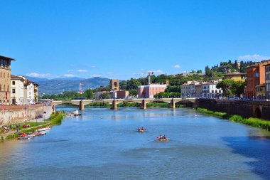 Floransa, İtalya, 11 Mayıs 2024: Açık mavi gökyüzü altında tarihi binalar ve köprüler bulunan Floransa 'nın arno nehrinin önü. Canlı manzara yeşil tepeleri ve uzak dağları kapsıyor, Floransa 'nın 