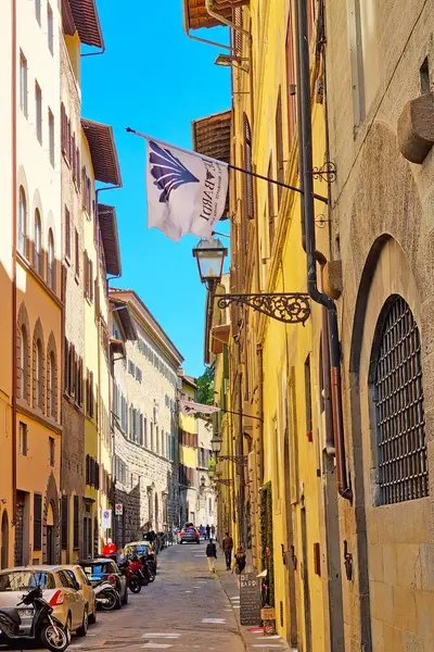 stock image Florence, Italia, May 11, 2024: Historic narrow street in florence, italy, featuring charming architectural facades with vibrant colors and flags. bicycles parked along the side, and distant pedestrians add to the urban atmosphere, capturing the esse