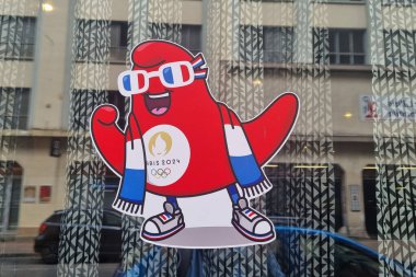 Lille, Fransa, 23 Haziran 2024: Paris 2024 Olimpiyat Olimpiyatları maskot çıkartması arka planda kentsel mimari yansımaları olan bir pencerede sergileniyor. 3D gözlük takan kırmızı ve beyaz maskot ve spor ayakkabıları heyecan ve coşkuyu simgeliyor. 