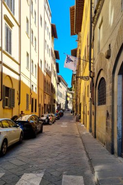 Florence, Italia, 11 Mayıs 2024: İtalya 'da çok iyi korunmuş tarihi yapılar sergilenen antika bir şehir sokak sahnesi. Arabalar kaldırım taşı sokağının iki tarafına park edilmiş ve bir binaya bayrak asılmış. Açık mavi gökyüzü t 'ye ekler