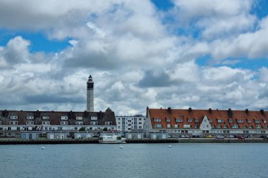 Calais, Fransa, 22 Haziran 2024: Dağınık bulutlarla dolu bir gökyüzünün altında, bitişik evlere sahip belirgin bir deniz feneri manzarası. Turizm promosyonları, mimari tasarım referansları ve kıyı yaşam tarzı temaları için ideal.