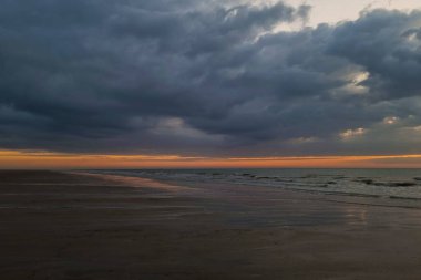 Uçsuz bucaksız bir sahilde büyüleyici bir günbatımı, fırtınalı, karanlık bulutlar, parlak, renkli ufka karşı dramatik bir tezat oluşturuyor. Islak kum çarpıcı gökyüzünü yansıtır, derinlik ve sükunet ekler.