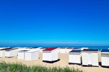 Beyaz plaj kulübeleri parlak mavi gökyüzünün altında kumlu bir plajda dizilmişti. Arka plandaki sakin okyanus sakin bir yaz tatili sahnesi yaratıyor. Yazı kutlayan posterler, seyahat broşürleri ve tebrik kartları için ideal..