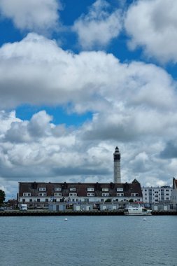Calais, Fransa, 22 Haziran 2024: Büyüleyici sahil kenti kıyı kenti kıyısı parçalı bulutlu bir gökyüzüne karşı ayakta duran tarihi bir deniz fenerine sahiptir. Sahne, sakin sulara yanaşmış teknelerle su kenarına dizilmiş tuhaf binaları içeriyor. ideal 