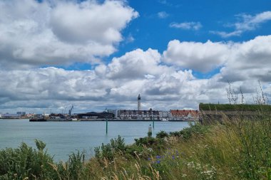 Calais, Fransa, 22 Haziran 2024: Deniz feneri ve kıyı şeridi boyunca manzaralı binaların yer aldığı bir kıyı kasabasının manzarası. Sahne yemyeşil otlar, kır çiçekleri ve dağınık bulutlarla dolu canlı bir mavi gökyüzü ile güçlenir. 