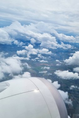 Pofuduk bulutların nefes kesici hava manzarası ve bir uçak penceresinden çekilen geniş manzaralar. Türbin motoru görünür durumda, seyahat ve havacılığın özünü vurguluyor. Gökyüzünün ve toprağın huzur dolu görüntüsü..