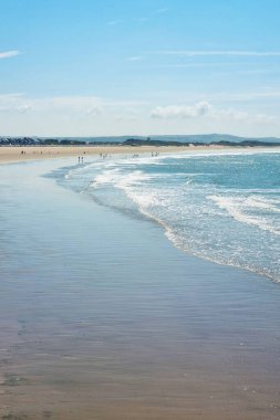 Berrak mavi gökyüzünün altında sakin dalgaların hafifçe dokunduğu geniş kumlu bir kıyı şeridine sahip sakin bir sahil manzarası. Uzakta, küçük insan grupları huzurlu kıyı ortamının tadını çıkarıyorlar, yaz tatili temaları veya seyahat tasarımları için mükemmel..