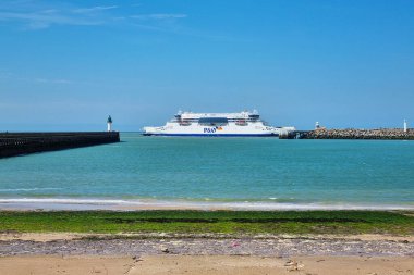 Calais, Fransa, 22 Haziran 2024: Sakin okyanus suları ve açık mavi gökyüzü tarafından çerçevelenmiş büyük bir feribotun deniz manzarası. Görüntü, liman girişlerini ve kıyı bölgesinin bir kısmını gösteriyor, sakin bir deniz atmosferini betimliyor.