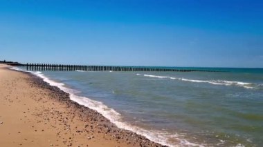 Sakin, turkuaz suya uzanan uzun bir ahşap iskelenin olduğu sakin bir sahil sahnesi. Yumuşak dalgalar kumlu kıyı şeridini çevreleyen çakıl taşlarıyla kaplı açık mavi gökyüzünün altında görünürde hiç bulut yok. Ortam huzurlu ve davetkar.