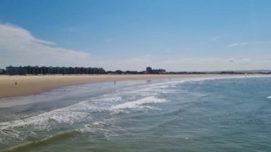 Sakin bir okyanus kıyısı sahnesi, yumuşak dalgalar, engin bir kumsal ve açık mavi gökyüzünün altındaki uzak binalar. Yaz tatili temaları ve baskı, kart veya poster gibi tasarım amaçları için mükemmel.