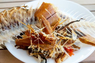 Parçalanmış balık kemikleri, çıtır çıtır deri ve beyaz bir tabakta dizilmiş balık pulları. Tabak, balıkların dokusunu ve kalıntılarını vurgular, gastronomik sunumlar ve besin odaklı sunumlar için uygun mutfak yaratıcılığını ve detaylarını vurgular.