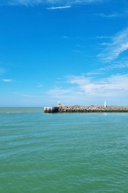 Berrak mavi gökyüzünün altında kayalık bir iskeleye tünemiş deniz feneri bulunan sakin bir deniz manzarası. Geniş okyanus ve canlı mavi su seyahat, dinlenme ve kıyı temalı tasarımlar için ideal bir durgunluk ve rahatlık hissi yaratır..