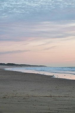 Gün batımında sakin bir sahile sahip sakin dalgalar, engin bir ufuk ve okyanusla birleşen pastel renkli bir gökyüzü. Huzurlu ortam rahatlama, seyahat ilhamı ve doğa hayranlığı için mükemmeldir..