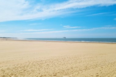 Sade bir sahil, altın rengi kumları olan sakin mavi bir denize doğru uzanır açık gökyüzünün altında ince beyaz bulutlarla süslenir. Uzakta bir gemi ufukta geziniyor ve birkaç insan yaz tatili temaları için ideal olan huzurlu kıyı şeridinin tadını çıkarıyor..